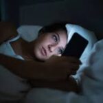 Seseorang menatap layar ponsel sebelum tidur di kamar gelap, menggambarkan kebiasaan digital malam hari yang bisa mengganggu kualitas istirahat.